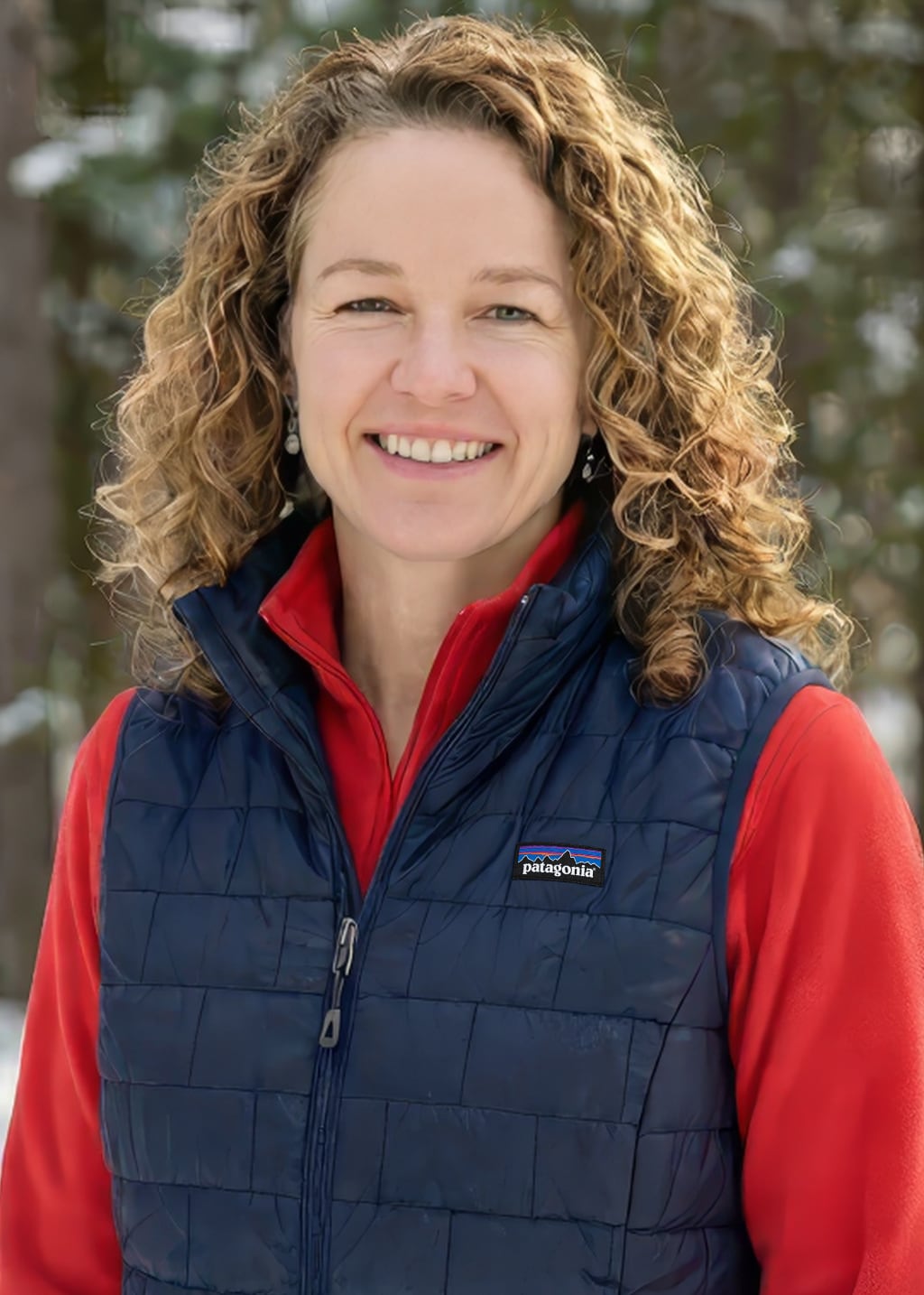 A woman with curly blonde hair smiles outdoors, wearing a red shirt and a navy blue Patagonia vest, with a snowy background and trees behind her.