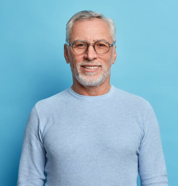 Older man with gray hair and beard, wearing glasses and a light blue sweater, smiling while standing against a plain light blue background.