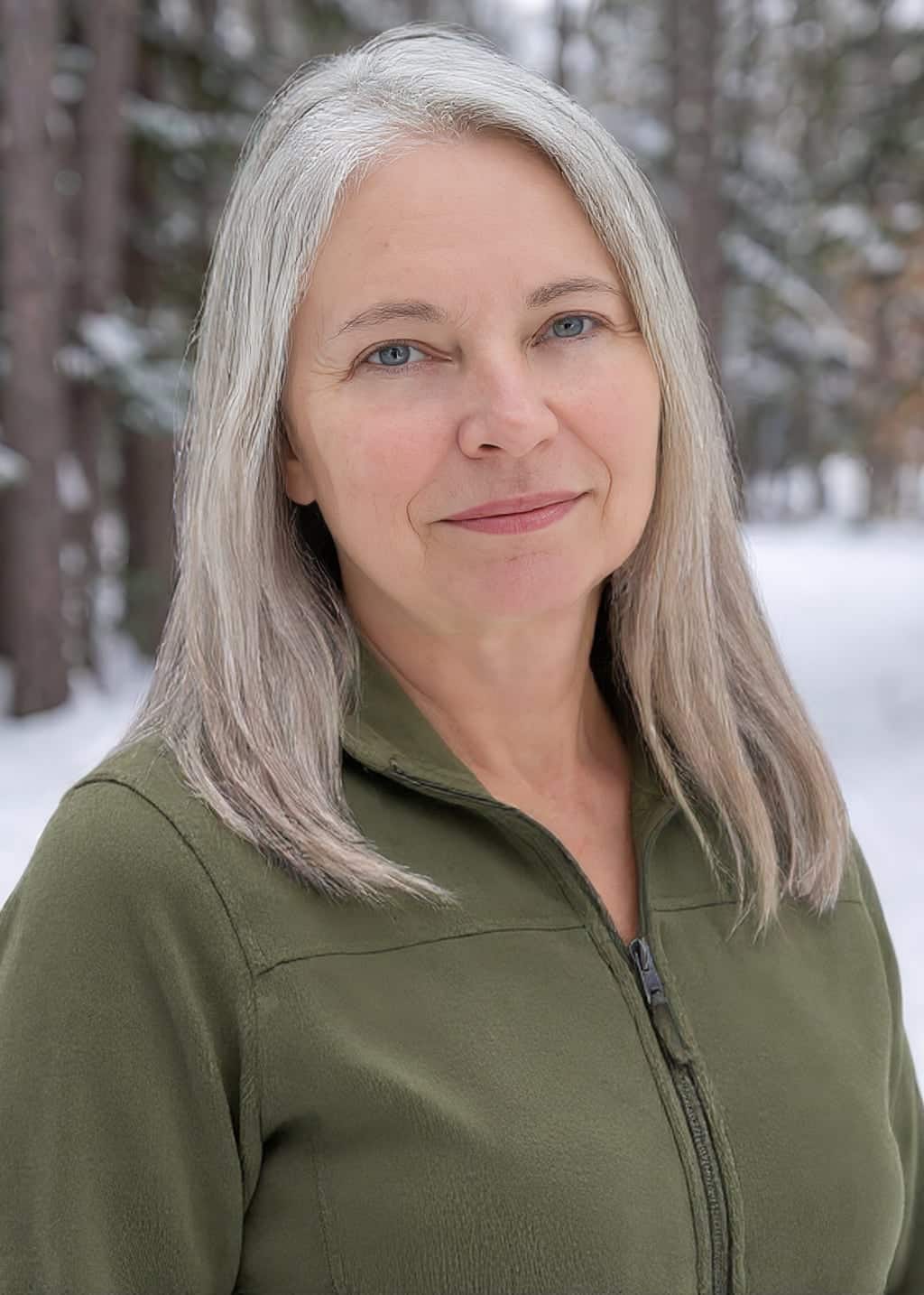 A woman with straight gray hair and blue eyes, wearing a green zip-up jacket, stands outdoors in a snowy forest, softly smiling at the camera.