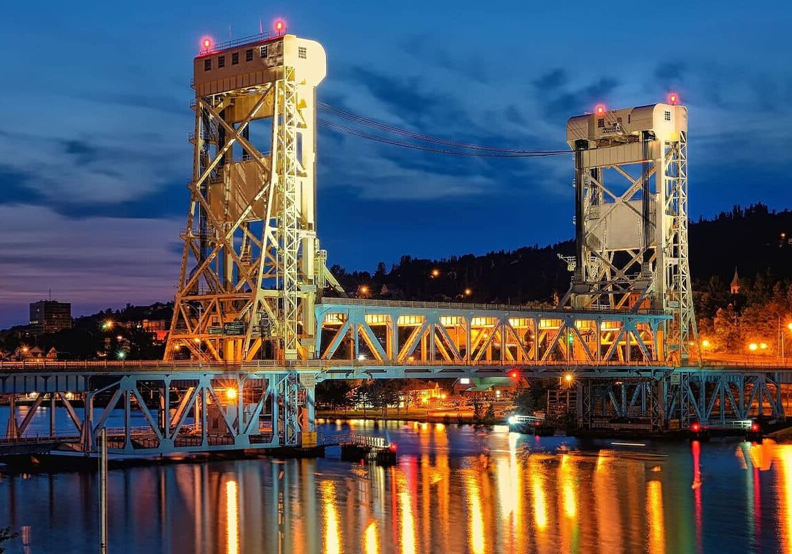 A brightly lit lift bridge spans a calm river at dusk, with its lights reflecting on the water and a twilight sky in the background. City lights and buildings are visible on both sides of the bridge.