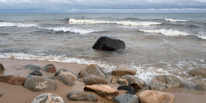 Rocky shoreline with various large stones on the sandy beach, gentle waves rolling in from a cloudy, distant horizon across the sea.